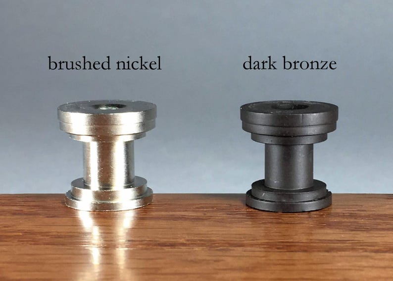 Pu&ograve; includere: Due componenti hardware cilindrici in metallo, uno con finitura in nichel spazzolato e uno in bronzo scuro. Ogni pezzo presenta una forma a bobina a gradini con parte superiore e inferiore piatte, etichettati come brushed nickel e dark bronze.
