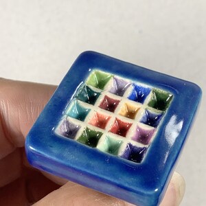 Puede incluir: Un azulejo de cer&aacute;mica cuadrado con un borde azul y un patr&oacute;n en relieve de cuadrados coloridos en el interior.