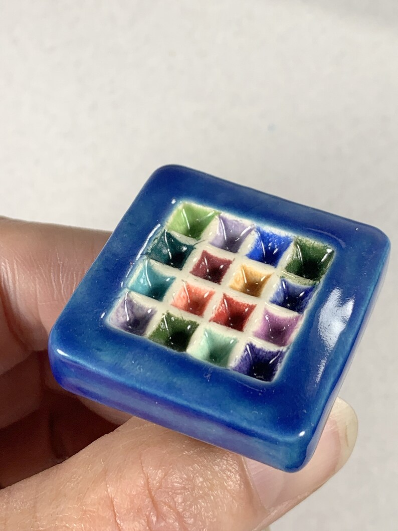 Puede incluir: Un azulejo de cer&aacute;mica cuadrado con un borde azul y un patr&oacute;n en relieve de cuadrados coloridos en el interior.