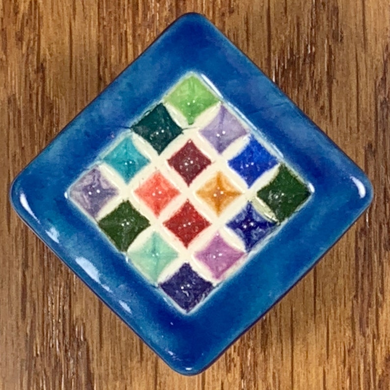 Puede incluir: Un azulejo cuadrado de cer&aacute;mica azul con fondo blanco y un patr&oacute;n de diamantes de colores. El azulejo tiene un acabado brillante y es perfecto para agregar un toque de color a cualquier espacio.