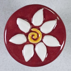 May include: A white ceramic flower with a yellow center on a burgundy background. The flower has six petals and a spiral design in the center.