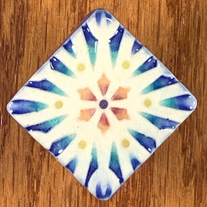 May include: A square ceramic tile with a floral design. The tile features a white background with a central flower in shades of orange and purple. Blue and teal accents radiate outwards, with small yellow dots. The tile is set against a wooden surface.