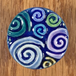May include: A round ceramic knob with a blue background and a swirling pattern of green, blue, purple, and white.