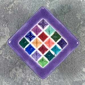 May include: A square ceramic tile with a purple border featuring a central grid of sixteen smaller diamond-shaped tiles in various colors including red, orange, yellow, green, blue, and violet, each with a raised center point.