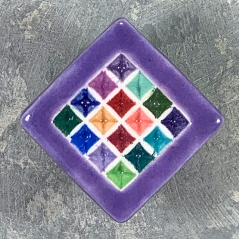 May include: A square ceramic tile with a purple border featuring a central grid of sixteen smaller diamond-shaped tiles in various colors including red, orange, yellow, green, blue, and violet, each with a raised center point.