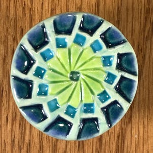 May include: A round, mosaic-style decorative piece with a light green center radiating outwards. The design incorporates teal, dark blue, and light blue square and trapezoid-shaped tiles. The piece is set against a wooden background.