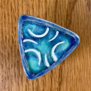 May include: A triangular ceramic dish with a blue and green glaze. The dish has a white design of curved lines.