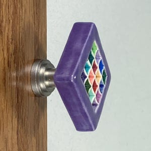May include: A diamond-shaped purple cabinet knob with a central grid pattern featuring small, recessed squares in shades of green, blue, red, and yellow. The knob is attached to a silver-toned metal base mounted on a wooden surface.