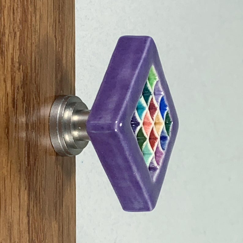 May include: A diamond-shaped purple cabinet knob with a central grid pattern featuring small, recessed squares in shades of green, blue, red, and yellow. The knob is attached to a silver-toned metal base mounted on a wooden surface.