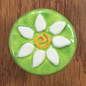 May include: A green ceramic knob with a white daisy flower design. The center of the flower has a yellow swirl.