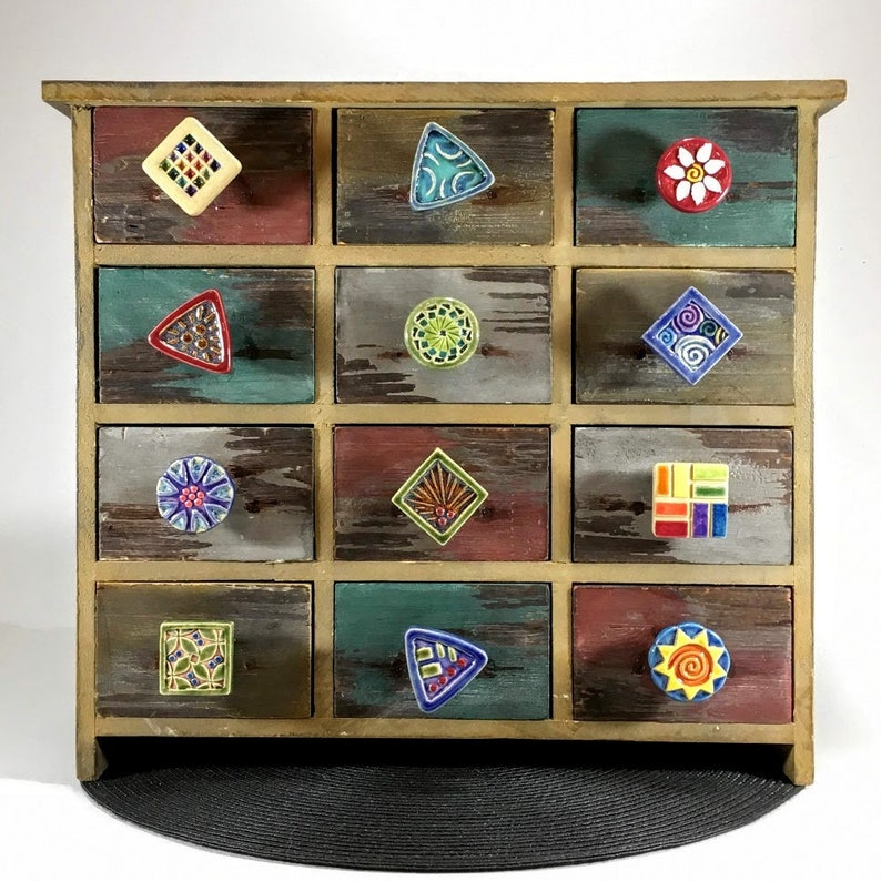 Puede incluir: Un armario de madera con 12 cajones, cada uno con una perilla de cer&aacute;mica &uacute;nica. Las perillas presentan varias formas geom&eacute;tricas y colores, incluyendo una flor, un sol, una estrella y un diamante.