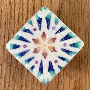 May include: A square, white ceramic tile with a symmetrical design. The design features a central flower shape in shades of orange and yellow, surrounded by blue and teal pointed shapes, and small purple dots. The tile is set against a wooden background.