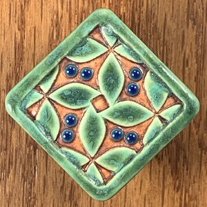 May include: A square ceramic tile with a diamond orientation, featuring a raised green leaf pattern on a terracotta-colored background. The design includes small, glossy, dark blue circular accents embedded within the leaf motifs.