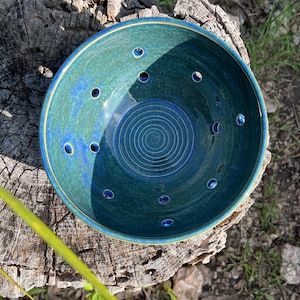 Könnte beinhalten: Handgefertigte Keramikschale mit türkisfarbener und blauer Glasur. Die Schale hat eine Reihe von Löchern am Rand und ein Spiralmuster in der Mitte. Sie steht auf einer verwitterten Holzoberfläche, ideal für die Küche.