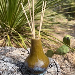 Può includere: Un piccolo vaso in ceramica bulboso con una smaltatura verde oliva lucida che sfuma in una base screziata blu e grigia. Cinque sottili bastoncini diffusori in legno chiaro sono inseriti nel collo stretto del vaso, rivolti verso l'alto.