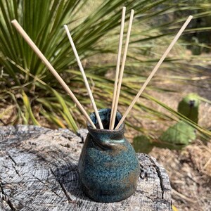 Puede incluir: Un pequeño difusor de cerámica hecho a mano con un esmalte moteado en tonos azul verdoso y marrón, con un cuello pellizcado y asimétrico. Cuatro varillas de madera clara están insertadas en la abertura, extendiéndose hacia afuera.