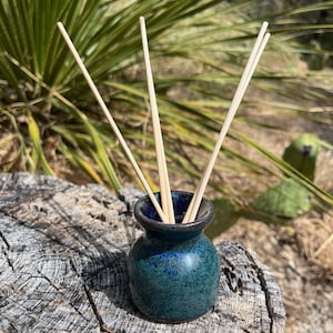 May include: A small, round, ceramic diffuser vessel with a glossy, mottled blue and green glaze. The neck of the vessel is narrow with a flared rim, and it contains four thin, light-colored wooden reeds extending upward from the opening.