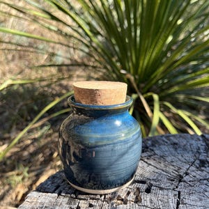 Puede incluir: Un pequeño tarro de cerámica hecho a mano con un tapón de corcho. El tarro es de color azul oscuro con vetas azules claras y blancas, y una base blanca. El corcho es de color marrón claro. El tarro está sobre una superficie de madera envejecida.