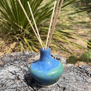 May include: A small, round, ceramic reed diffuser vessel with a glossy blue and green mottled glaze. The neck is narrow and flares slightly at the top, holding four thin, light-colored wooden reeds that extend upward from the opening.
