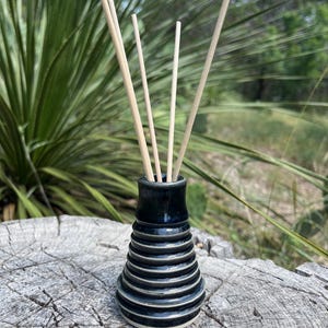 Puede incluir: Un difusor de aceites esenciales de cerámica negra con un diseño acanalado y varillas claras. El difusor está sobre una superficie de madera desgastada, con follaje verde en el fondo. Un artículo decorativo para el hogar.