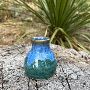 May include: A small, handcrafted ceramic vase with a rounded body and a narrow neck. The vase features a gradient of colors, transitioning from a deep teal green at the base to a vibrant blue and a gold rim. The vase is sitting on a weathered wooden surface.