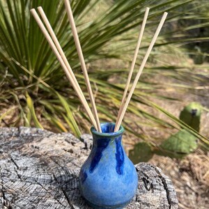 May include: A small, bulbous ceramic vase with a glossy finish in shades of blue and green, featuring a narrow neck and a wide base. Four thin, light-colored wooden reeds are inserted into the opening of the vase, extending upwards.