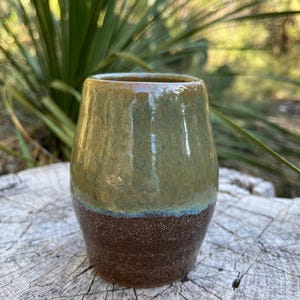May include: A small, green and brown ceramic mug with a speckled glaze. The mug is sitting on a wooden surface.