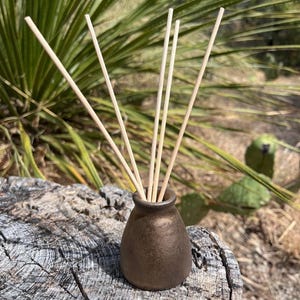 May include: A small, tapered, metallic bronze-colored ceramic diffuser vessel with a narrow neck. Four thin, light-colored wooden reed sticks are inserted into the opening of the vessel, extending upwards in a fan-like arrangement.