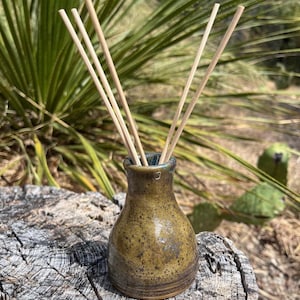 May include: A small, bulbous ceramic vase with a mottled olive-green and brown glaze, featuring a narrow neck and a small stamped mark near the rim. Four thin, light-colored wooden reed diffuser sticks are inserted into the opening of the vase.