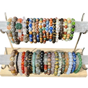 May include: A two-tiered display stand holds numerous beaded bracelets. The bracelets feature a variety of colors and materials, including gemstones and wooden beads. The stand has a light wood base and silver metal supports.