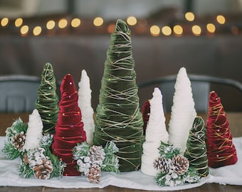 May include: A set of seven miniature Christmas trees made of fabric and decorated with gold and red ribbon. The trees are arranged on a white tablecloth with pine sprigs and pinecones.