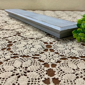 May include: A light blue wooden shelf with a distressed finish. The shelf is sitting on a white crocheted tablecloth.