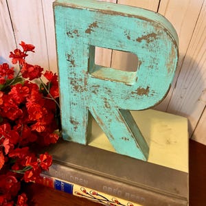 Pu&ograve; includere: Una lettera R in legno di colore blu turchese con una finitura usurata. La lettera &egrave; appoggiata su una pila di libri con il testo "Children" e "Greg Read" visibile.
