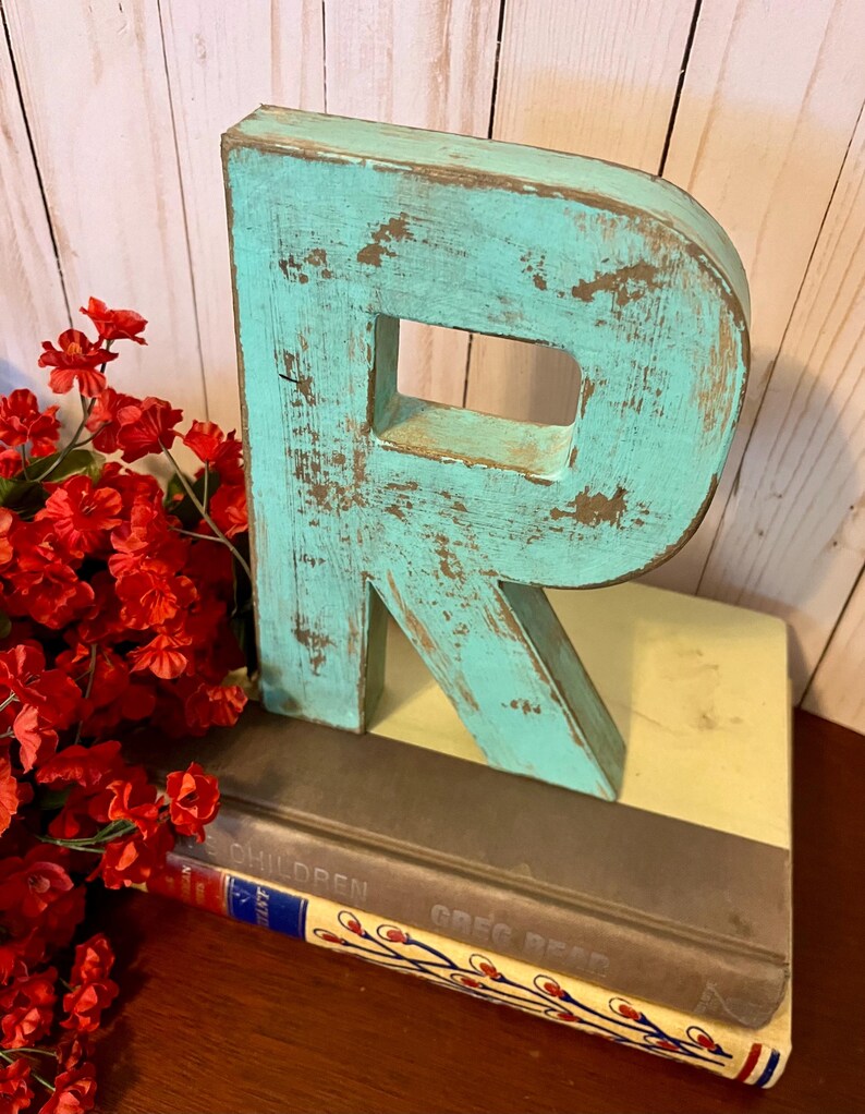 Pu&ograve; includere: Una lettera R in legno di colore blu turchese con una finitura usurata. La lettera &egrave; appoggiata su una pila di libri con il testo "Children" e "Greg Read" visibile.