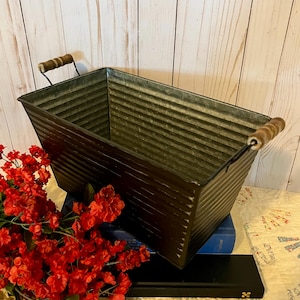 May include: A rectangular, dark metal planter with a corrugated design and wooden handles. The planter is filled with red flowers and sits on top of books. The handles are made of wood and are attached to the metal frame.