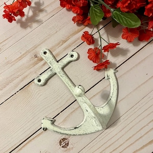 May include: A distressed white metal anchor wall hook. The anchor has a crossbar and a curved lower portion. Red flowers are in the background. The anchor is approximately 7 inches long.