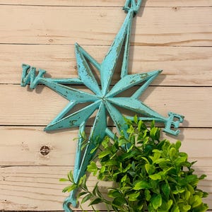 Peut inclure: Une étoile de boussole en métal bleu turquoise avec les directions N, S, E et W. La boussole est montée sur un fond en bois blanc avec une plante verte au premier plan.