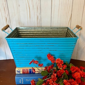 以下が含まれることがあります： 木製の取っ手が付いた、ターコイズブルーの金属製のプランター。プランターは、赤い花が前に置かれた本の束の上に置かれています。