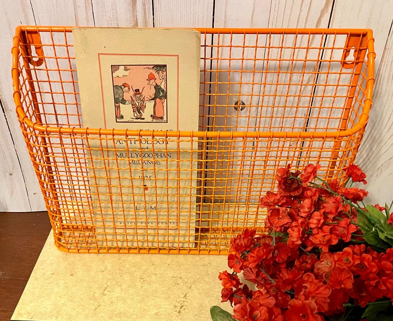 K&ouml;nnte beinhalten: Orangefarbener Wandhalter aus Metalldraht mit einem Buch mit dem Titel "Anthology of Mollycoddle Melange" im Inneren. Der Halter ist an einer wei&szlig;en Holzwand montiert, im Vordergrund befinden sich rote Blumen.