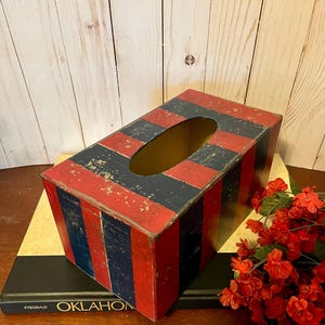 May include: Rectangular wooden tissue box with a distressed red and navy blue striped pattern. The box has an oval opening on top and is sitting on a book. Decorative floral arrangement in the background.