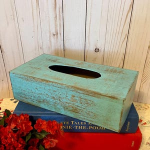 May include: A rectangular tissue box with a distressed turquoise finish and a long oval opening. The box sits atop two books, one blue and one red, with a bouquet of red flowers in the foreground.