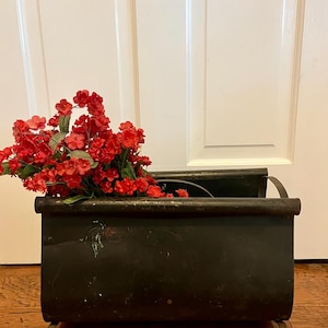 Puede incluir: Un revistero de metal negro con flores rojas en su interior. El revistero tiene una forma curva y se encuentra sobre un suelo de madera.