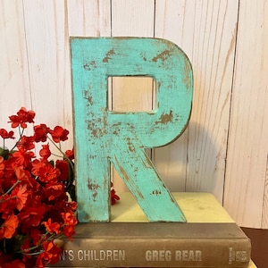 Pu&ograve; includere: Una lettera R in legno di colore blu turchese con una finitura usurata. La lettera &egrave; appoggiata su una pila di due libri. Il libro superiore ha il titolo "Greg Bear" e il libro inferiore ha il titolo "The American Poets".