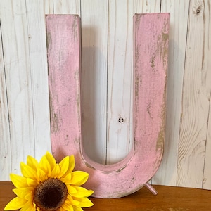 Peut inclure: Une grande lettre U en bois rose avec une finition vieillie. La lettre est posée sur une surface en bois avec un tournesol jaune devant elle.