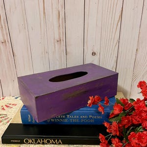 May include: A rectangular, purple wooden tissue box with an oval opening on top. The box has a distressed finish, and is resting on top of a stack of books. Red flowers are in the foreground.