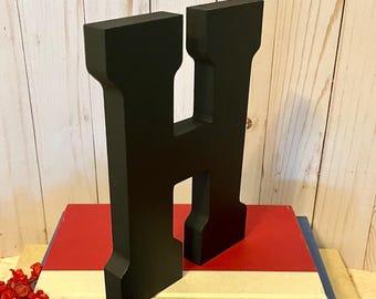 May include: A large, black wooden letter "H" stands on a stack of books with red, white, and blue covers. The books are adorned with red flowers. The letter is a bold, decorative element.