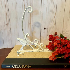 Puede incluir: Un gancho de pared de metal blanco envejecido con forma de pájaro, con un brazo curvo y adornos decorativos. El gancho está montado sobre un libro titulado "Oklahoma" con flores rojas en el fondo.