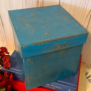 May include: A square, lidded box with a distressed, bright blue painted finish. The box has visible wear along the edges and surfaces, revealing a lighter base layer beneath the blue paint. The lid is flat and fits over the top of the box.