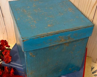 May include: A square, lidded box with a distressed, bright blue painted finish. The box has visible wear along the edges and surfaces, revealing a lighter base layer beneath the blue paint. The lid is flat and fits over the top of the box.
