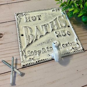 Pode incluir: Um sinal de ferro fundido branco com um gancho, apresentando o texto "HOT BATHS 25c" com um visual desgastado e vintage. O sinal tem uma borda decorativa e um gancho para pendurar.
